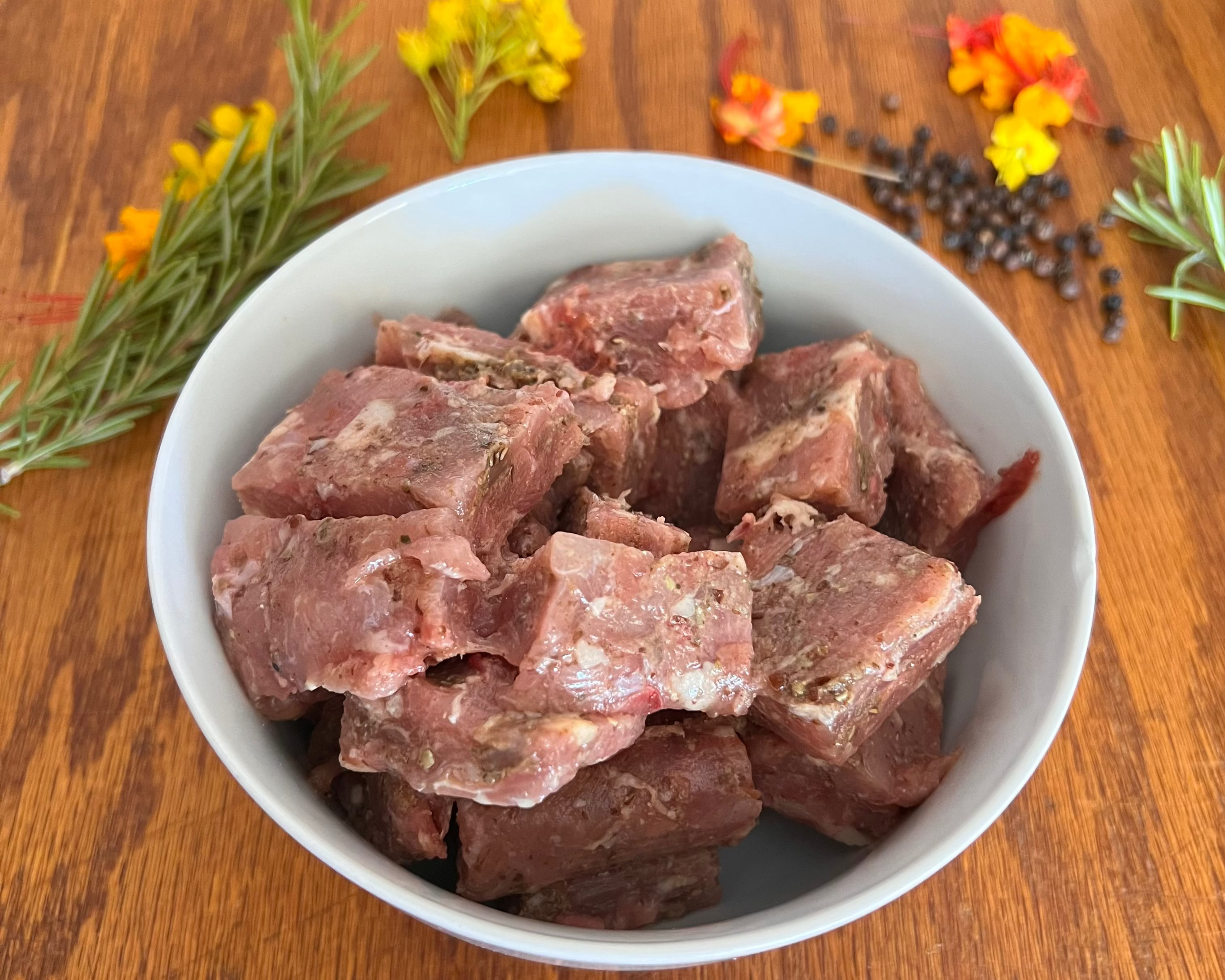 Raw meat chunks in a white bowl on a wooden surface with herbs and spices.